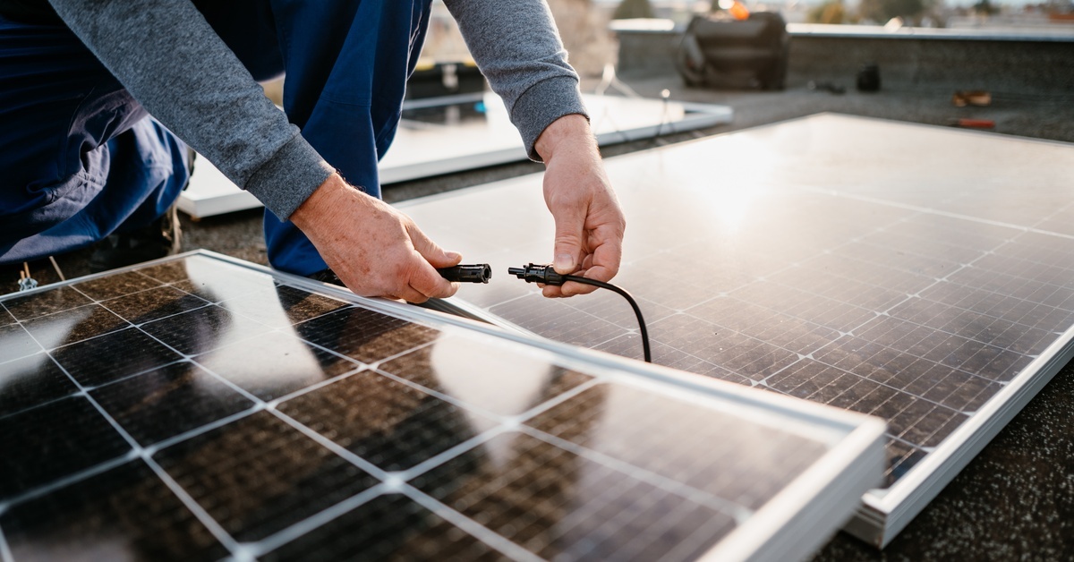 A worker installing solar energy panels on a rooftop. They are connecting to panels to each other with wiring.