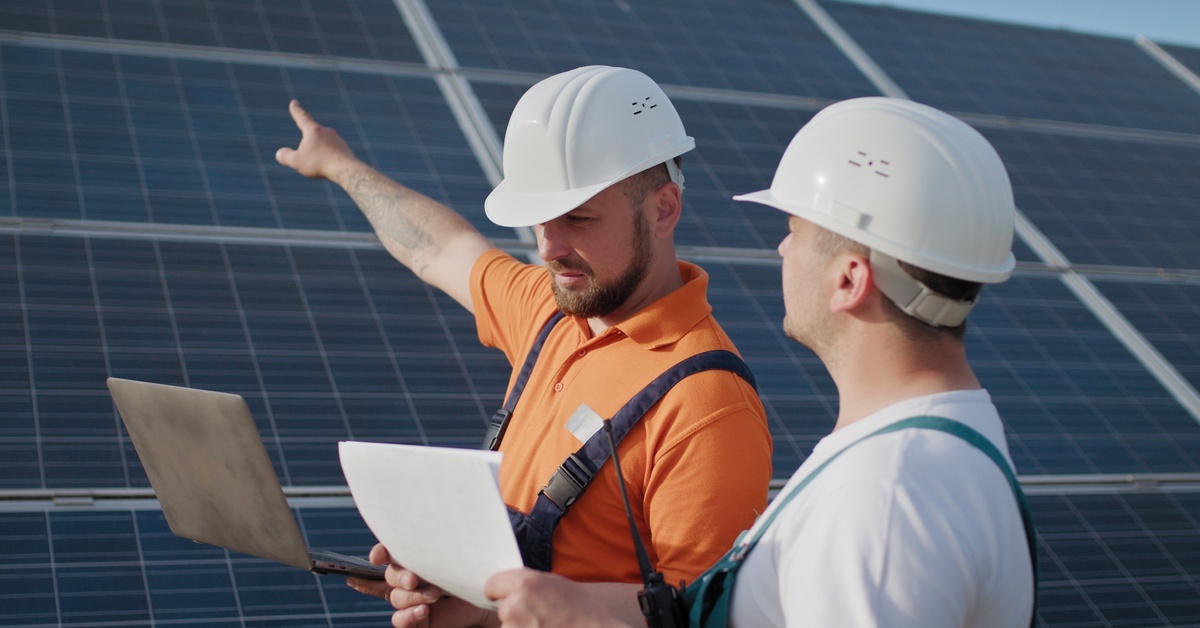 Two engineers wearing hard hats and carrying laptops are reviewing a solar project. One of them is pointing at a panel. Two engineers wearing hard hats and carrying laptops are reviewing a solar project. One of them is pointing at a panel.