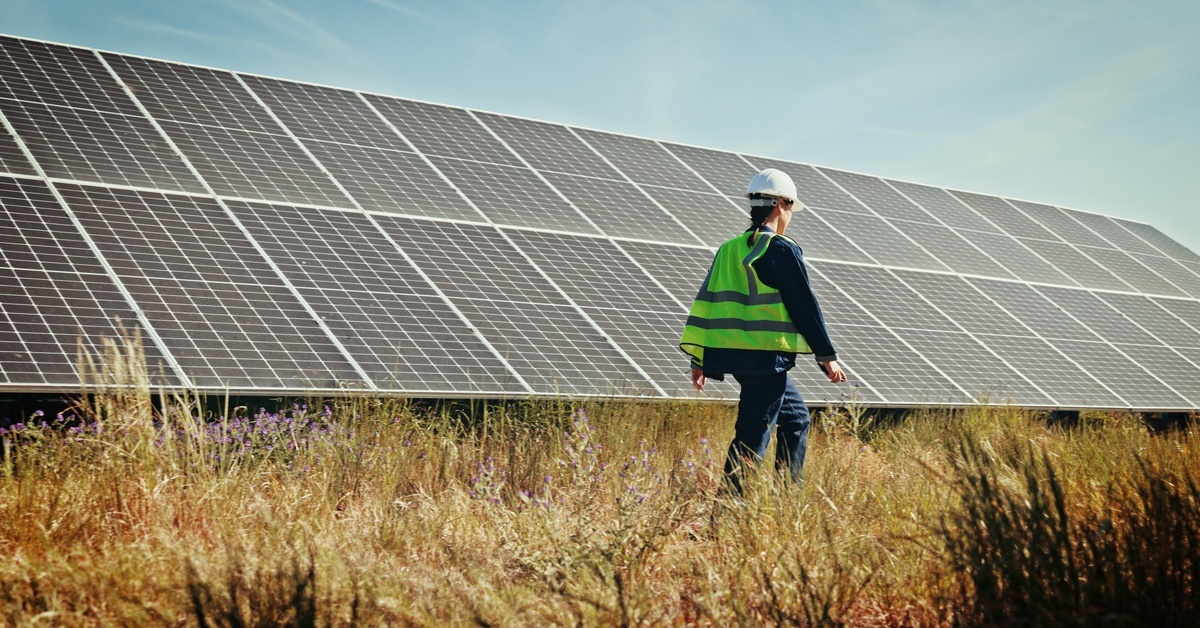 A worker in safety gear walking through a field with a solar panel installation. She is inspecting the project. A worker in safety gear walking through a field with a solar panel installation. She is inspecting the project.