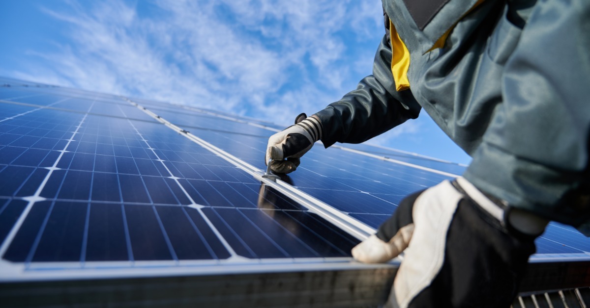 A person wearing work gloves installing a solar panel beneath a bright blue sky with scattered white clouds.