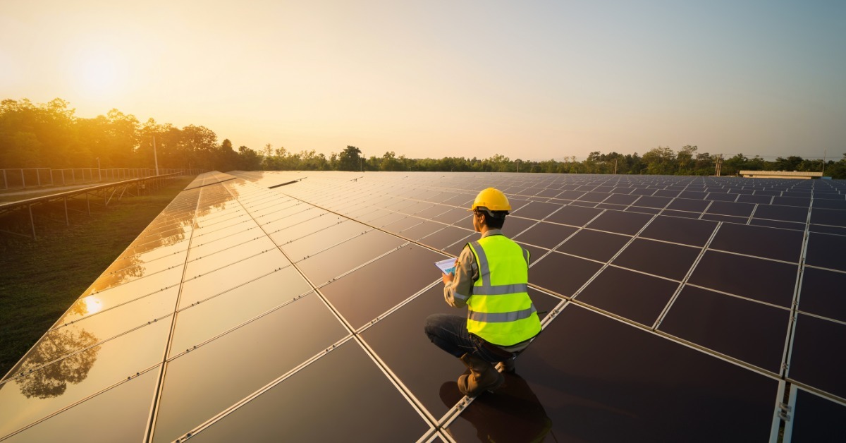 A person in a yellow hard hat and safety vest crouching on solar panels while holding a tablet at sunset.