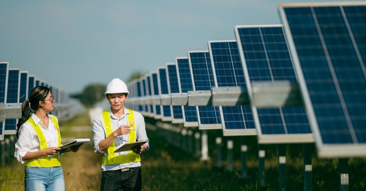 Two engineers wearing safety vests walking between rows of solar panels while holding clipboards and a tablet.