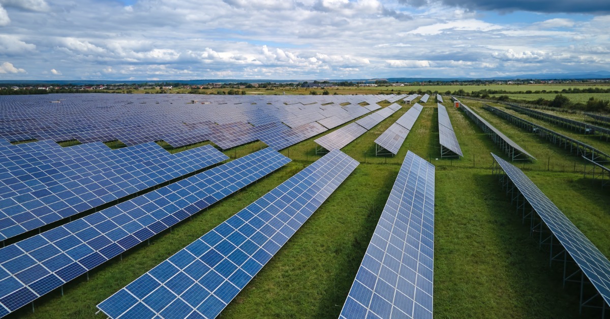 A large solar farm with rows of blue panels on green grass beneath a cloudy blue sky and distant fields.