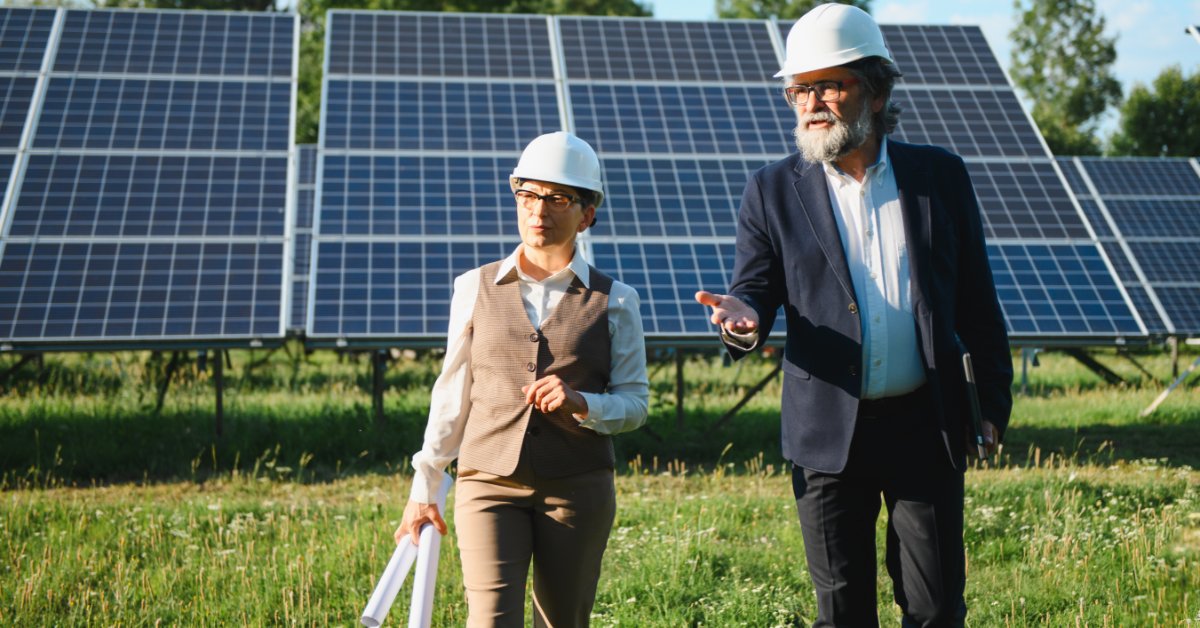 Choosing Between Engraved and Printed Solar Labels Two people wearing hard hats and safety vests reviewing papers beside rows of solar panels in a grassy field.